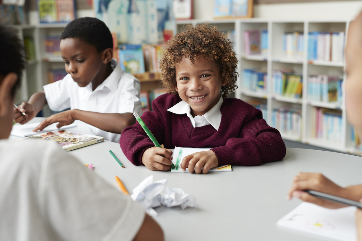 Primary children in class, curly haired boy writing and looking at camera