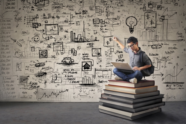 A man is sitting on a pile of books and looking at a laptop