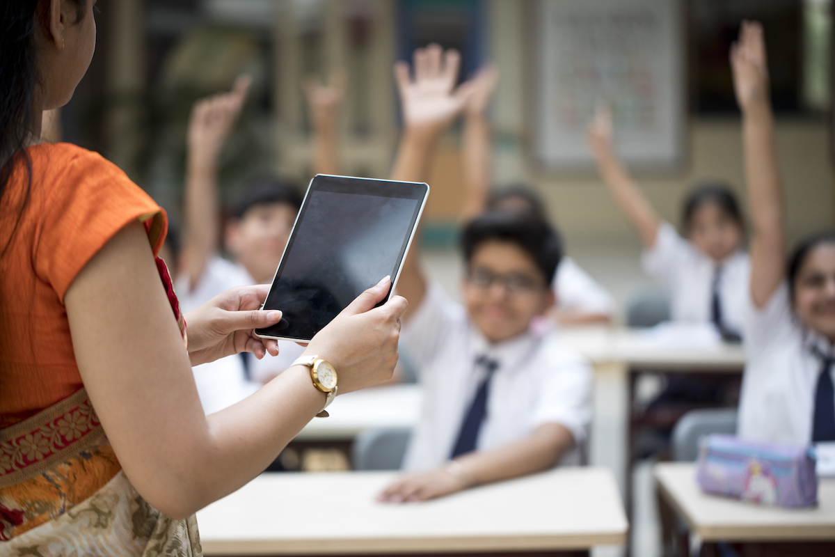 Teacher seen from behind, students with hands up ready to answer