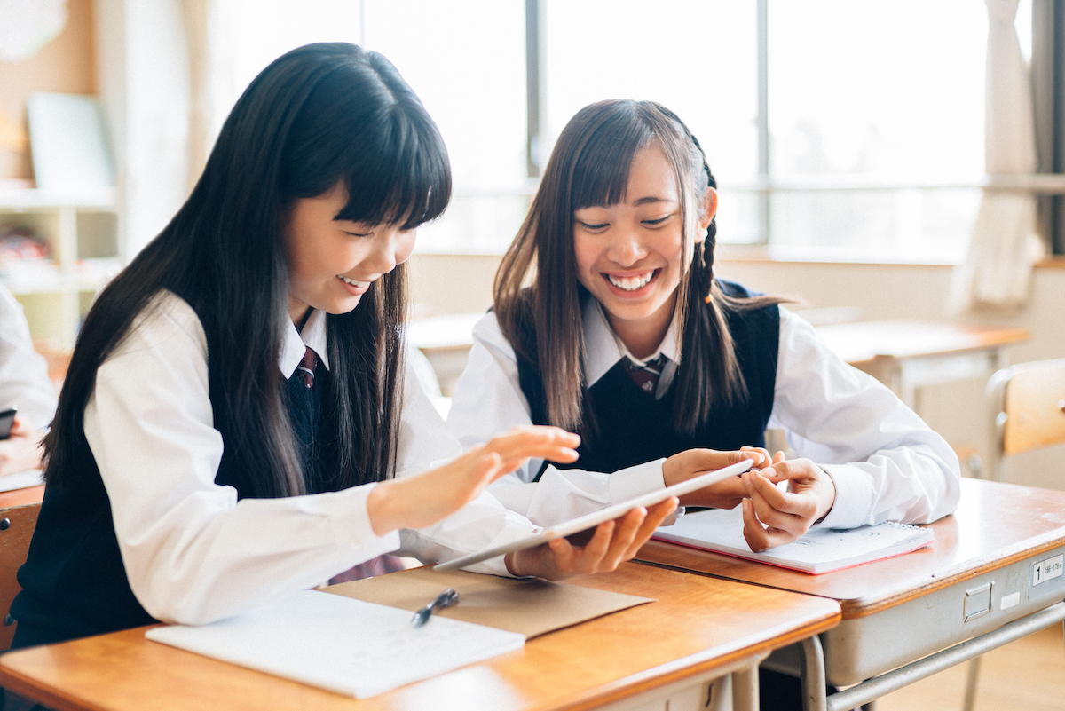 Two female Japanese school students in a classroom sitting down looking at a tablet device