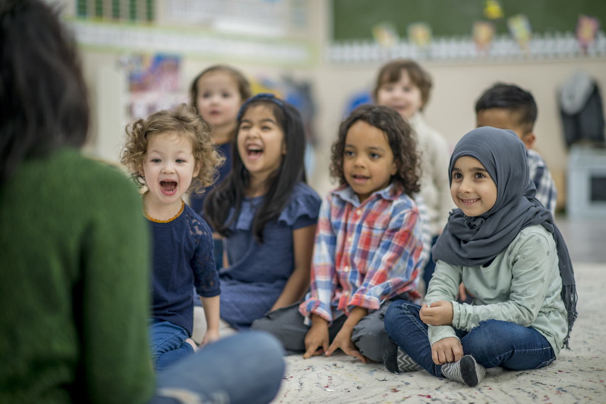 children sitting on the floor, listening to a teacher, also sitting