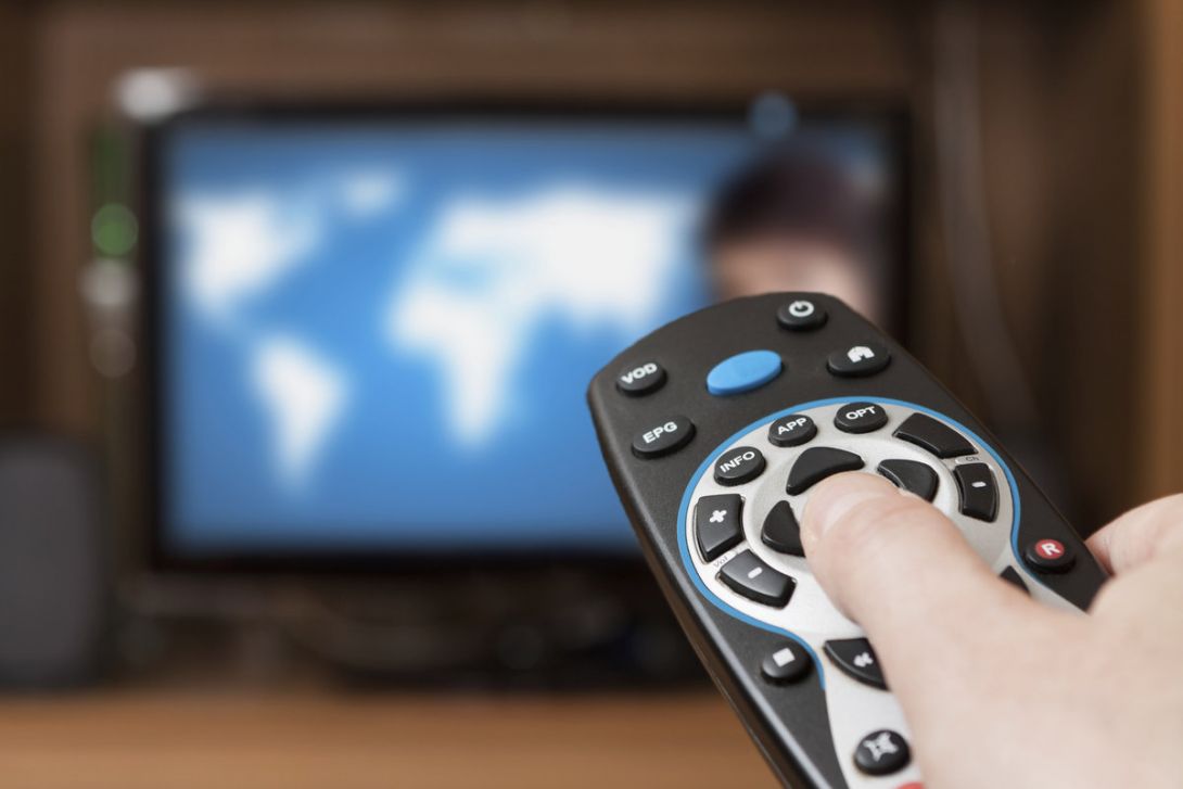 A hand is holding a TV remote, pointed at a TV in the background