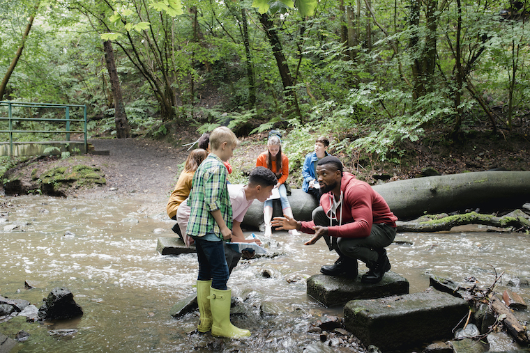 A teacher on a rock in a shallow river explaining something to his primary students