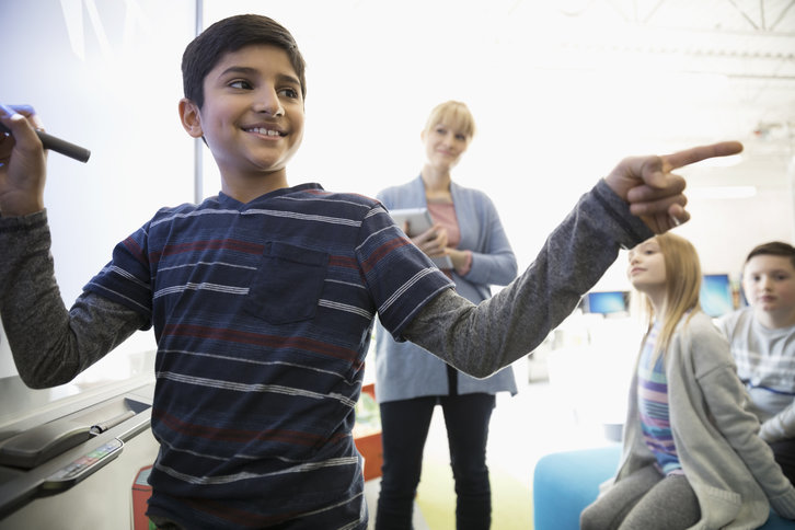 Young student standing at a whiteboard pointing at classmate