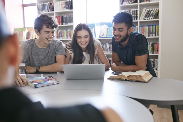 group of young students in a library looking at a laptop together