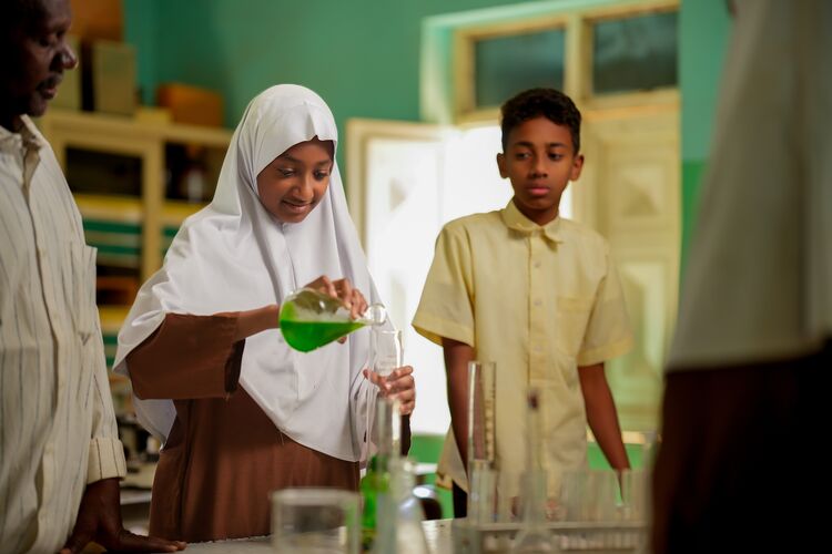 Female teenage student pouring green liquid into chemistry flask in a classroom.  